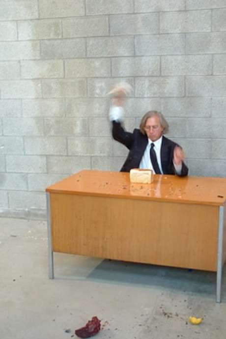 Jimmie Durham sits behind a desk in a suit, performing a bureaucratic ritual of crushing objects. A visitor presents a prehistoric stone tool that shatters Durham, yet he remains impassive, stamps a receipt and hands it back as the visitor exits. The sequence repeats throughout the 92‑minute runtime, a minimalist commentary on authority and violence.